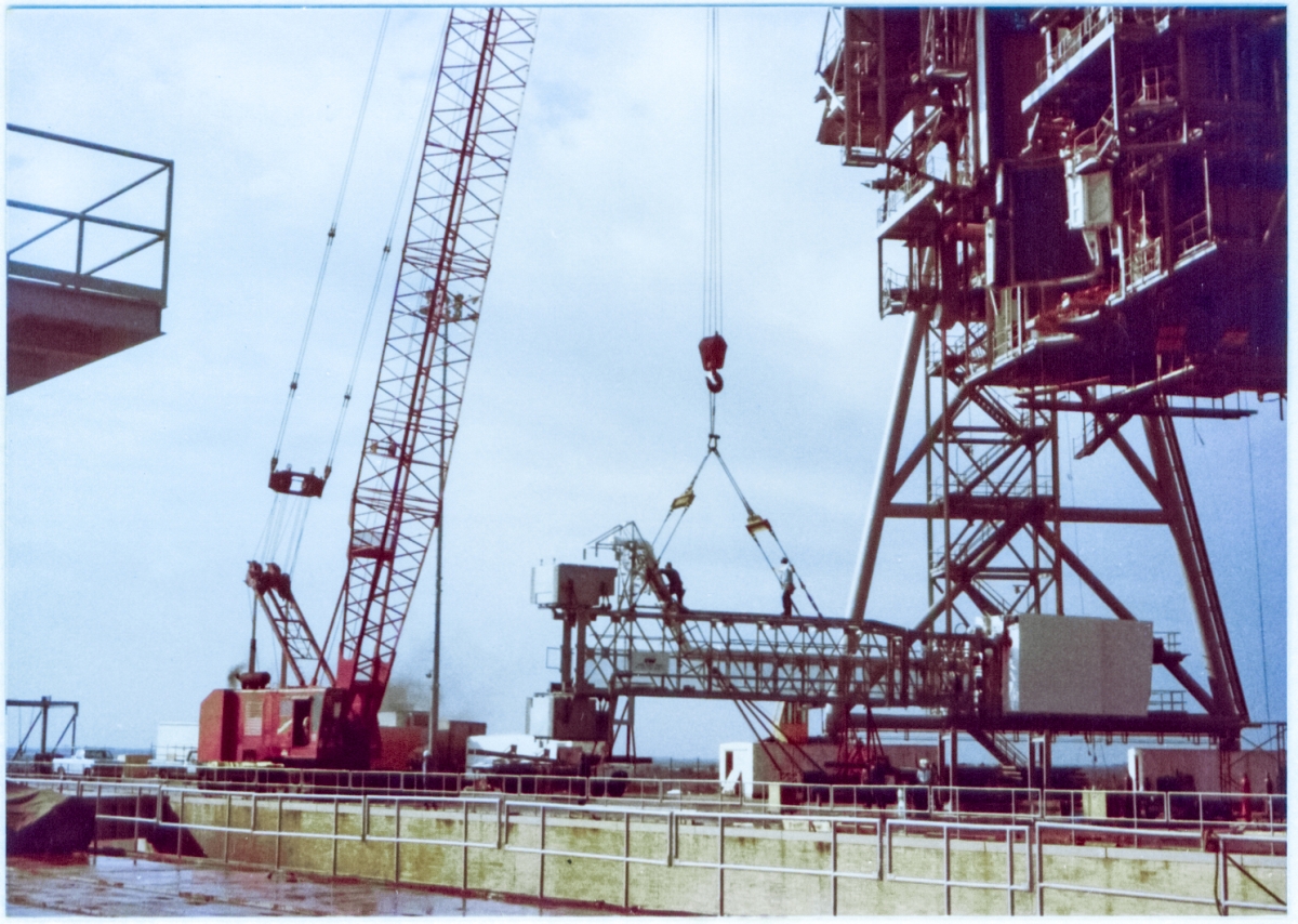 The Orbiter Access Arm at Space Shuttle Launch Complex 39-B, Kennedy Space Center, Florida, has been lifted off of the ground and is being reoriented, and a pair of union ironworkers from Local 808 are riding the suspended load, verifying all is well with the lifting gear and making such adjustments with it as necessary, prior to commencing with the main lift, which will end with the OAA securely fastened to the Fixed Service Structure between the 200' and 220' elevation framing steel levels. Of note in this image is the fact you can see that the support stanchions which hold the arm up when it is resting on solid ground have been lifted into the air along with the arm itself. Those stanchions were firmly attached to the arm truss, and were not removed until the lift proceeded in earnest.